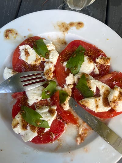 A white plate of caprese salad, featuring sliced tomatoes, fresh mozzarella, and basil leaves drizzled with a balsamic glaze and whole grain mustard, rests on a dark wooden table outdoors. A fork is positioned to the left of the plate, and a knife is to the right. A partial view of a wine glass filled with white wine is visible at the top of the image. The scene appears to be at a restaurant in Bièvres, France.