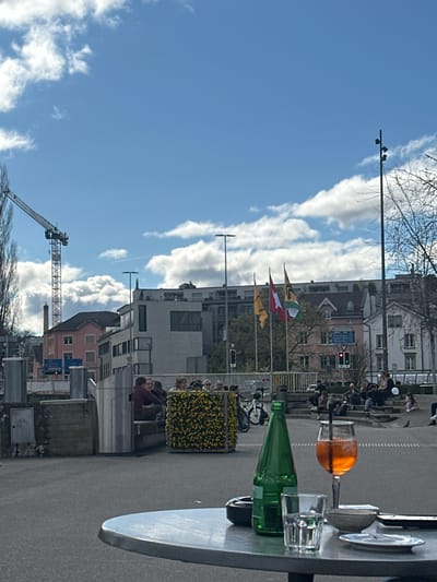 The image captures an outdoor scene in Schaffhausen, Switzerland, with a focus on a table setting in the foreground. On the table, a green glass bottle lies horizontally, with a filled wine glass containing an orange liquid (likely a spritz) and an empty drinking glass beside it. Silverware and plates are also present.

The background reveals a street scene with pedestrians, including people gathered in groups. Flags (likely Swiss and cantonal) are visible among buildings of various architectural styles. A construction crane is present in the distance. The sky is blue with scattered white clouds. Street signs and a traffic light can also be observed. The scene conveys a vibrant outdoor social gathering in an urban environment.