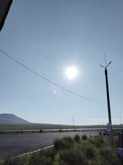 The image depicts an outdoor scene on a clear day with a bright sun. A power line stretches diagonally across the sky from the left side of the frame towards the right. On the right side, a tall, dark utility pole with multiple crossbars and what appears to be a lightning rod at the top is prominent. There is a white sign attached to the pole.
In the foreground, a paved surface, likely a road or parking lot, is visible, bordered by green grass and shrubbery. In the mid-ground, a flat, grassy landscape stretches towards a gently sloping hill in the background. The sky is a pale blue and appears cloudless, with the sun positioned in the upper left quadrant, creating a lens flare effect. The overall impression is of an open, rural or semi-rural environment. Based on the provided metadata, this scene is located in Московское, Russia.