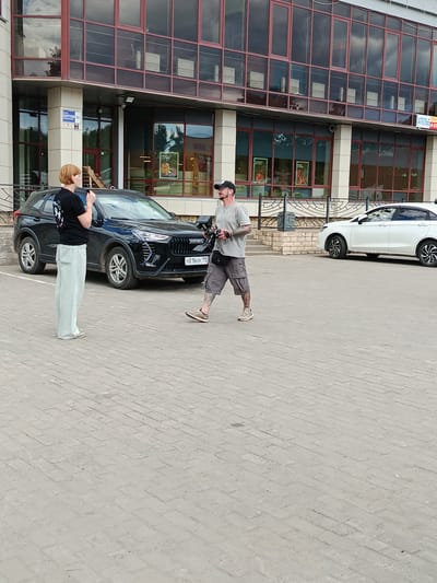 In Koltushi, Russia, two individuals stand on a paved lot in front of a modern building with large glass windows. A woman with short red hair, wearing a black t-shirt and light blue wide-leg pants, stands near a black SUV. She holds a phone or similar device to her ear. A man with tattoos on his arms and legs, wearing a gray t-shirt, shorts, and a black baseball cap, walks past the SUV holding a camera. He has earbuds in his ears and a black pouch around his waist. A white sedan is parked to the right of the black SUV. The building has a sign that reads "АТЕЛЬЕ" and "КОПЕЙКИ" on it. The weather appears to be overcast.
