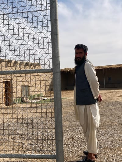 This image, captured in Nakhonay, Afghanistan, depicts an outdoor scene featuring an adult male positioned to the right of a galvanized chain-link fence. The man wears a light-colored shalwar kameez and a dark, sleeveless vest, with dark slip-on shoes. He has a full dark beard and dark hair, and is looking directly towards the viewer with his hands clasped behind his back. The chain-link fence, with a prominent vertical metal post, occupies the left and central portions of the foreground and midground. Behind the fence, the ground consists of dry dirt and gravel. In the background, two traditional mud-brick or adobe-style buildings are visible. The building on the left features a wooden door and a flat roof. Near this structure, green and white striped mats or textiles are laid on the ground. A second, partially obscured building with a thatched roofline is located further back to the right. The sky overhead is partly cloudy, displaying patches of blue.