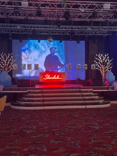 The image depicts an indoor stage setup, likely for a performance. In the background, a large screen displays a dynamic visual of a person playing a guitar, superimposed over a snowy, abstract landscape. The screen is framed by stage lighting rigs and rigging. In the foreground, a raised stage area is visible with a neon sign spelling out "Shambala" in red script. To the left and right of the stage, decorative trees with illuminated accents stand. Several speakers are positioned on the stage. In front of the stage, tiered seating with a dark, glossy finish is visible. The floor is covered in a patterned red carpet. The scene is lit by a combination of stage lights and the illuminated decorations.