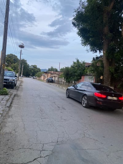 The image depicts a street scene in Miriantsi, Bulgaria. A dark-colored car is driving down the street, with a stork perched in a nest on a utility pole to the left. The street is paved with cracked asphalt and lined with houses and trees. The sky is cloudy.