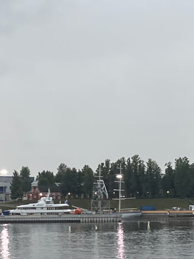 A white motor yacht is docked at a harbor on a cloudy day. The yacht is long and sleek, with multiple decks and windows. It is moored alongside a pier that is lined with bollards and a walkway. Behind the yacht, there is a line of trees and buildings. In the water, the reflection of the yacht and the sky can be seen. The image is taken from across the water, looking towards the docked yacht. This scene is consistent with a harbor in the city of Imeni Sverdlova, Russia, which is known for its waterfront and maritime activities.