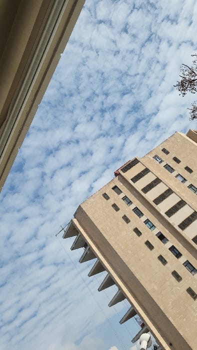 The scene presents an upward-angled view of urban architecture against a sky in Magaluf, Spain. The primary subject is a multi-story building, oriented diagonally from the bottom-right towards the upper-left. Its facade is constructed of light tan masonry units, featuring multiple rectangular windows and a distinctive series of triangular projections or balconies along its left-facing side. Horizontal dark-slatted vents are integrated into upper floor sections, and a segment of a red rooftop is visible at its apex. A second, lighter-toned building is partially visible along the extreme left edge of the frame, showing a vertical segment of its facade with a lighter trim. The sky above is light blue, extensively covered with patchy, white altocumulus-like clouds. Fine, dark linear elements, consistent with utility wires, extend diagonally across the mid-ground. Bare tree branches are visible against the sky in the upper-right corner. A white, cylindrical object with a rounded cap is partially present at the bottom-right.