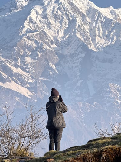A person dressed in a black coat and beanie stands on a mossy outcrop, with snow-capped mountains forming the backdrop. The location is Lwang Ghalel, Nepal. The individual is facing toward the mountain range, the perspective suggesting they are gazing out at the scenery. The branch of a small tree is visible next to the person.