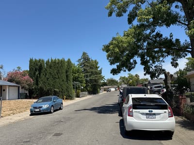 A residential street in North Highlands, California, is depicted on a clear, sunny day. The street is paved with asphalt and slopes slightly downhill from right to left. On the left side of the street, a blue Volkswagen Eos is parked on the asphalt adjacent to a sidewalk. Behind the Volkswagen, a single-story house with a light-colored exterior and a white garage door is visible, with a trash bin nearby. The yard in front of the house is dry and covered in brown grass. A line of tall, dark green cypress trees runs along the property line. Further down the street on the left, there is a pink flowering bush.
On the right side of the street, a white Toyota Prius is parked, facing away from the viewer. It has a license plate that reads "6WY1951". Behind the Prius, a black Jeep Wrangler is parked. Further down the street, several other vehicles are visible, including a dark gray SUV and a white car. The right side of the street is lined with houses, fences, and trees, with a large deciduous tree with green leaves prominently positioned in the foreground, partially obscuring the sky. The sky is a clear, bright blue with no clouds. Shadows are cast across the street, indicating the position of the sun.