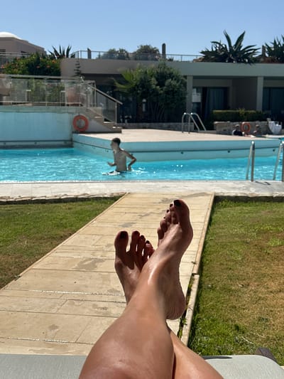 A first-person perspective shot shows a person's bare feet with dark nail polish resting on a light-colored stone pathway. Beyond the pathway is a well-maintained green lawn, and further back is a swimming pool filled with blue water. A young boy is visible in the pool. In the background, there are buildings with balconies, lush green plants, and trees. The sky is clear and blue, indicating a sunny day. The overall scene depicts a relaxing outdoor setting, likely a resort or hotel in Tavronitis, Greece, given the vegetation and architecture.