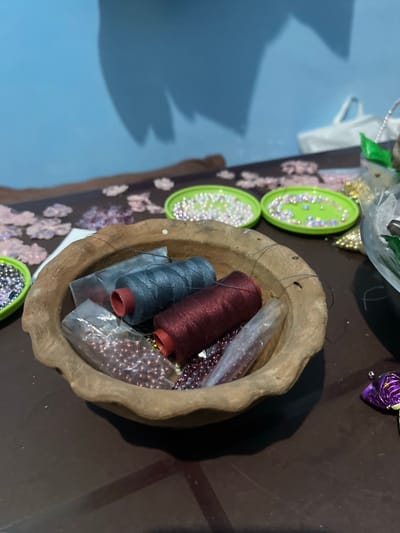 The image depicts a close-up of a crafting table, showcasing various materials for beadwork and sewing. In the foreground, a rustic, earthy-toned bowl contains spools of thread, one a deep burgundy and the other a dark grey, along with several small plastic bags filled with beads. The beads appear to be primarily purple and metallic brown, with some clear or iridescent ones also visible. A needle and thread are casually placed across the bowl. To the left of the bowl, a green dish is filled with small, shimmering beads, some appearing pearl-like and others iridescent. Further to the left, another green dish holds similar beads. In the background, a light blue wall casts a shadow of what appears to be a large, abstract shape. The table surface is a dark brown, with some scattered pink and metallic decorative elements, possibly sequins or small floral cutouts, visible in the mid-ground. To the right, a metallic purple object, possibly a candy wrapper or decorative item, rests on the table. The scene suggests an indoor setting where crafting activities are taking place. This scene is not located in Bandarbireuen, Indonesia, as there are no visual elements that indicate this specific location.