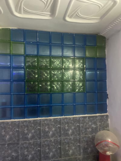 The image depicts a wall composed of blue and green glass blocks, set above a tiled lower section of the wall. The glass blocks are arranged in a grid pattern, with the green blocks forming a central horizontal band. Above the glass block wall, there is a white, textured ceiling. The lower portion of the wall is covered in dark, speckled tiles. In the bottom right corner of the image, a red object with a clear, rounded top is visible, possibly a dispenser or a container, with what appears to be a white bird inside. The setting appears to be an interior space, possibly a bathroom or a similar utility area. The image does not provide any specific visual cues that can be used to pinpoint the location within the city of Kporva, Nigeria.