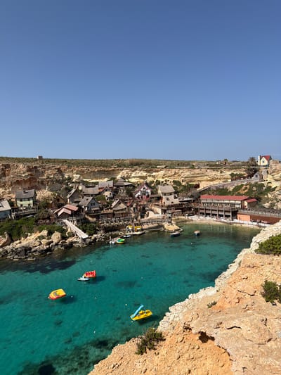 An aerial or high-angle view captures a picturesque coastal scene, likely Popeye Village in Mellieħa, Malta, under a clear blue sky. The image depicts a small, rustic village nestled into a steep, rugged cliffside that descends towards a tranquil turquoise bay.
The village, composed primarily of wooden buildings with varying roof colors (some red, some grey), appears built directly into and along the base of the rocky cliffs. Green vegetation and sparse trees dot the landscape around the structures. People can be seen scattered throughout the village and on a small beach area, enjoying the setting.
The calm, clear teal-blue water of the bay is vibrant and inviting, revealing parts of the seabed. Several colorful recreational boats and watercraft are dotted across the surface: a prominent yellow and blue pedal boat, a red and yellow pedal boat, and another yellow boat. A white paddleboard is also visible near a red boat. A few individuals are in the water or on these boats, engaging in leisure activities.
The right side of the image features a dramatic, sun-drenched rocky cliff face that sharply drops into the sea, providing a natural boundary to the bay. The bright, cloudless sky and strong sunlight suggest it is midday or early afternoon, indicating pleasant and warm weather. The scene evokes a sense of leisure and vacation. No discernible text is visible in the frame.
