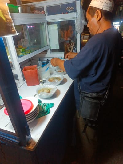 A man wearing a white cap and a navy blue shirt is standing behind a food stall in Luwuk, Indonesia. The stall appears to be outdoors, with a white countertop and a glass display case. Several bowls of food, likely bakso or meatballs, are arranged on the counter. A red plastic container and a stack of white plates with a pink lid are also visible. The man is preparing food, and his reflection can be seen in the glass case. A black fanny pack is worn around his waist. The overall scene suggests a busy street food environment.