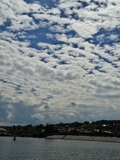 The image depicts a waterfront scene in Verhnij Uslon, Russia, characterized by a clear blue sky filled with fluffy white clouds on the left side and a more overcast, streaky cloud formation on the right. A body of water, likely a river, occupies the right side of the frame, its surface rippled by a gentle breeze. Along the embankment, a line of buildings and dense green trees suggests a residential or small town area. The architecture of the buildings appears varied, with some roofs visible and hints of various colors.
The perspective suggests the photo was taken from a boat or the water's edge, looking towards the shore. The lighting indicates it is daytime, with the sun likely high in the sky, casting soft shadows. The clouds suggest fair weather with potential for some cloud cover as the day progresses. The overall mood is peaceful and serene, with a sense of calm by the water. There is no visible text in the image. The scene captures a moment of natural beauty and tranquil village life in a Russian setting.