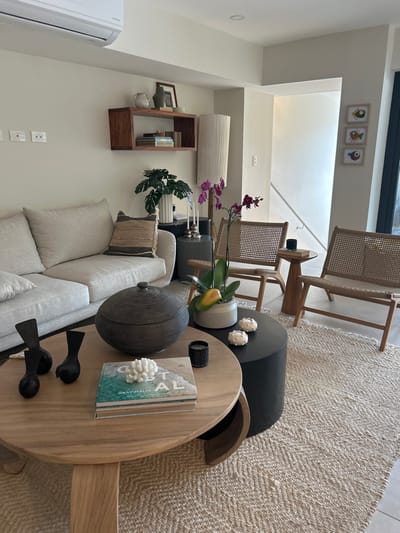 The image depicts a living room located in a private residence in Playa Herradura, Costa Rica, specifically at the coordinates 9.658 latitude and -84.673 longitude. The room features a white couch with beige cushions, a wooden coffee table adorned with a decorative bowl, a book, a coral ornament, and two black bird sculptures. Two wicker chairs are strategically positioned in the room, with a wooden end table between them. An orchid plant in a ceramic pot is situated on the floor near the wicker chairs. Three framed artworks depicting colorful fish are hung on the wall adjacent to a doorway leading to another room. A wooden bookshelf is mounted on the wall, showcasing decorative objects and books. The room is characterized by a light and airy ambiance, with beige walls and a beige rug covering the floor. The room is well-lit, with natural light streaming in from the doorway. The scene is devoid of any human presence.