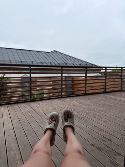 A person's legs, wearing beige crocs with charms, are stretched out on a wooden deck. The deck is made of parallel wooden planks. In the background, a modern house with a dark metal roof is visible. A wooden fence with horizontal slats runs along the edge of the deck. The sky is overcast. The image appears to have been taken from a seated or reclining position. The location is identified as Semiluki, Russia.