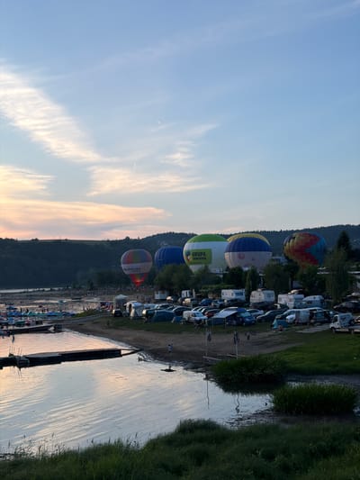 This image depicts a lively scene at a lakefront campground in Gródek nad Dunajcem, Poland, during what appears to be an outdoor event, possibly a hot air balloon festival. In the foreground, calm lake waters reflect the soft hues of the twilight sky, with a wooden dock jutting into the water. The shoreline is dotted with numerous tents and camper vans, indicating a camping or festival setup. A variety of vehicles, including cars and vans, are parked near the tents.

Further back, the lake is bustling with activity. Numerous small boats, including pedal boats and kayaks, are scattered across the water, with many people visible enjoying leisurely activities. A crowd of people is gathered on the shoreline, observing the events.

Dominating the scene are several colorful hot air balloons, some of which are inflated and positioned on the ground, while others appear to be preparing for or have just completed a flight. One prominent balloon features the text "GRUPA PINGWINA" and "MAŁOPOLSKA ZAGRODA". The sky above displays a soft gradient of blues and pastels, with wispy clouds and faint contrails from aircraft. The background is characterized by a lush, dark green forested hillside. The overall atmosphere is one of a festive outdoor gathering by the water.