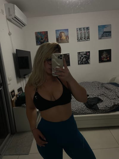 A mirror image captures an individual with long blonde hair, wearing a black Gymshark-branded sports bra and teal leggings. The individual holds a light-colored smartphone with triple cameras, obscuring the face, and is wearing eyeglasses. A thin bracelet is present on the right wrist. The scene is an interior room in a private residence located in Yavne, Israel. The background features a white wall with six square-format art prints arranged in a grid; identifiable prints include Travis Scott's 'Astroworld' album cover. A white split-unit air conditioner is mounted high on the left wall, beneath which a black flat-screen television is affixed. To the right, a white bed frame is visible, covered by a grey and white geometric patterned duvet, with a darker garment resting on it. A white cabinet with framed items is situated to the left of the bed. The flooring consists of light-colored tiles, and a grey floor mat is positioned near a sliding glass door on the far left of the frame.