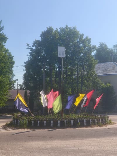 The image captures a view of a roundabout in Kady, Russia, adorned with a circular arrangement of brightly colored flags. The flags, predominantly in hues of yellow, blue, pink, and red, are attached to poles and are fluttering in the breeze. In the center of the roundabout, a tall pole stands topped with a white, rectangular structure, possibly an antenna or a light fixture. The roundabout is surrounded by a decorative black and white metal fence. Beyond the roundabout, lush green trees form a backdrop, and a building with a grey roof is visible in the distance. The ground is paved with asphalt, showing some wear and tear. The sky above is clear and blue, suggesting a sunny day. There are no people or vehicles present in the immediate vicinity of the roundabout. The overall scene depicts a public space decorated for an event or as a permanent feature of the town's landscape.