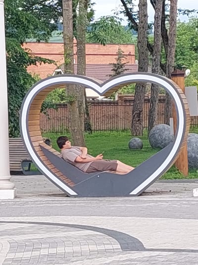 A person is reclined on a heart-shaped bench in what appears to be a park in Alikonovka, Russia. The bench is made of wood and metal, with a white trim around the heart shape. The person, dressed in a t-shirt and shorts, is holding a phone in their hand. In the background, there are trees, a brick building, a fence, and large decorative spheres. The ground is paved with cobblestones.