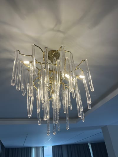 The image displays a close-up, low-angle shot of a modern chandelier hanging from a white ceiling. The chandelier features a gold-colored, branch-like structure from which numerous clear, cylindrical glass elements are suspended, resembling icicles. These glass pieces are illuminated by embedded lights, creating a warm glow and casting interesting shadows on the ceiling. The background is a blend of the white ceiling and a darker, bluish wall or ceiling feature below, with a hint of a window and curtains visible at the bottom. The scene appears to be an interior residential or commercial space. No specific location in Moni Community, Cyprus is identifiable from the image itself.