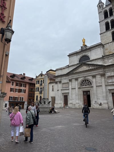 In the city of Annecy, France, a wide, cobblestone square is dominated by the imposing facade of a large, stone church. The church features a grand entrance with double wooden doors, arched windows, and ornate architectural details. A golden statue of a robed figure, likely religious, stands atop the church's roofline. To the left of the church, a cluster of colorful, multi-story buildings with traditional shutters line the square. A tall, obelisk-like monument stands in the mid-ground, flanked by statues.

Several people are present in the square. In the foreground, a woman in a pink sweatshirt and white pants walks away from the camera, carrying shopping bags. Beside her, a man in a green jacket and dark jeans also carries shopping bags. Further back, a person on a bicycle rides away from the viewer towards the church. Other individuals are scattered throughout the square, some sitting near the monument and others walking in various directions. The sky is overcast, and the overall atmosphere suggests a moderately busy day in a historic European city. A streetlamp is mounted on a building to the far left, near a sign with red lettering.
