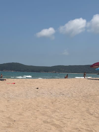 The image depicts a beach scene with a clear blue sky and scattered white clouds. The foreground is dominated by sandy beach, with some shells and small pebbles visible. In the background, the turquoise sea meets the shore with gentle waves. A densely forested hill forms the backdrop to the beach.
Several people are visible on the beach and in the shallow water. Near the water's edge, a person appears to be standing on a surfboard. Further back, a woman in a bikini is walking towards the water, and another person is partially visible in the sea. In the distance, other beachgoers and kite surfers can be seen. A red kite is lying on the sand.
The sun is shining brightly, suggesting it is daytime, likely midday or early afternoon. The weather appears to be warm and pleasant, ideal for a beach outing. The overall atmosphere is one of leisure and recreation. The location appears to be a coastal area, with the visual cues of the sea, sand, and hills consistent with a beach resort. No text is visible in the image.