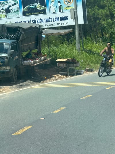 The image depicts a roadside scene in Thôn 6, Vietnam. In the foreground, a grey asphalt road with yellow lane markings is visible. To the left of the road, a dark green truck is parked, its bed covered by a dark tarp. The truck appears to be loaded with various items, including baskets of what seem to be fruits and vegetables. Next to the truck, a makeshift stand is set up with more baskets and some stacked cardboard boxes. In the middle ground, a person wearing a light brown jacket and sunglasses is riding a motorcycle. Behind the roadside stall and the motorcycle, dense green vegetation and trees form the background, suggesting a rural or semi-rural environment on the outskirts of a city. A billboard with text in Vietnamese is partially visible above the roadside stall.
