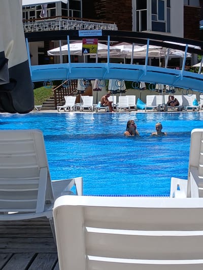 This image captures a sunny day at a swimming pool, likely within the complex of the Aquapark in Sapareva Banya, Bulgaria. The pool is a vibrant blue with a textured bottom. Two young women are visible in the water; one is nearer to the camera and has her hands near her face, while the other is further back and looking forward. In the background, several white lounge chairs are arranged around the pool, some unoccupied and others with people sunbathing. One person sits on a chair near the edge of the pool, reading. Above the pool area, a white bridge-like structure spans across, adorned with blue railings and hanging circular decorations. Beyond the bridge, the facade of a building with multiple windows and balconies is visible, suggesting it is part of the resort or hotel complex. The overall scene depicts a typical leisure environment, with clear water and a relaxed atmosphere.