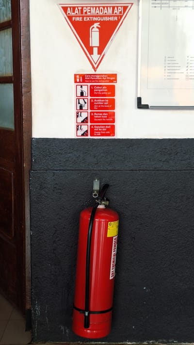 The image depicts a fire safety setup on a two-toned wall, likely indoors. On the left, a white wall displays a red triangular sign labeled "ALAT PEMADAM API FIRE EXTINGUISHER," indicating a fire extinguisher location. Beside it, a set of instructions (in Indonesian and English) outlines the steps for using a fire extinguisher. The instructions include pulling the safety pin, aiming at the base of the fire, squeezing the handle, and sweeping from side to side.
To the right, a black textured wall holds a red "DRY CHEMICAL POWDER" fire extinguisher. The extinguisher is secured to the wall with black straps. The lower part of the frame shows a dark brown wooden structure, possibly a counter or a wall panel. The text 'KEDUNG GEDER' indicates the location in the city of Karawang, Indonesia.