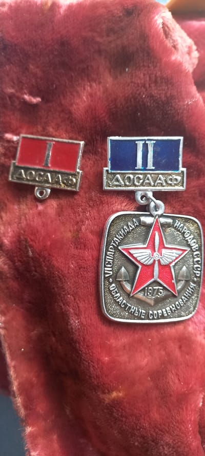 The image displays three Soviet-era badges laid out on a red, plush fabric surface. The badges are arranged from left to right, with the largest, a medal, positioned at the bottom right.
The leftmost badge is rectangular with a red top section and a silver bottom section bearing the inscription "ДОСААФ". A small loop is visible at the bottom of this badge.
The middle badge is also rectangular, with a blue top section and a silver bottom section, also inscribed with "ДОСААФ". A loop is present at the bottom, and this badge is attached to a larger medal below it via a chain.
The largest object is a square-shaped medal with rounded corners. It features a red, five-pointed star in the center with a propeller motif. The star is superimposed on a silver background. Around the star, a silver border contains inscriptions in Cyrillic. The top section reads "VIICPARTAKHAAAH". The bottom section reads "ОБЛАСТНЫЕ СОРЕВНОВАНИЯ" and "1975". There are also two anchor symbols visible on the left and right sides of the central star.
The plush red fabric appears to be a piece of cloth or a garment, possibly velvet or a similar material, with a textured surface. The lighting in the image suggests an indoor setting, but no specific environmental context such as windows, furniture, or the city of Novopavlovka, Kyrgyzstan, is depicted or inferable from the image itself. The image focuses solely on the badges and the fabric they are placed upon.