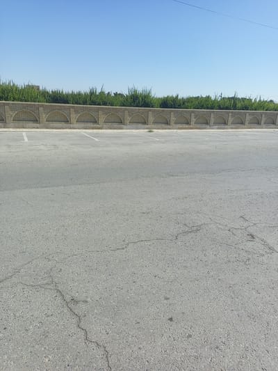 The image depicts an outdoor scene in Shar-Bakr, Uzbekistan, on a clear day. In the foreground, a cracked asphalt parking lot is visible, with faint white parking lines. A decorative brick wall with arched openings runs along the background. Behind the wall, a dense line of green trees forms a visual barrier. The sky above is a clear, bright blue. The scene appears to be devoid of any people or vehicles.