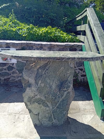 In the city of Sobótka, Poland, an outdoor scene features a table with a flat, polished, green stone top supported by a large, rough, grey stone base. The table is positioned on a paved area with flagstones, partially in shadow. To the right of the table, a weathered green wooden staircase with railings ascends. Behind the table, a stone wall is visible, topped by lush green foliage and vegetation. A sign with Polish text, reading "ZAPRASZAMY", meaning "We invite you", is mounted on the stone wall to the left of the table. Another smaller sign with an illustration and Polish text is partially visible beneath the table's stone top. The environment appears to be a park or a historical site, bathed in bright sunlight.