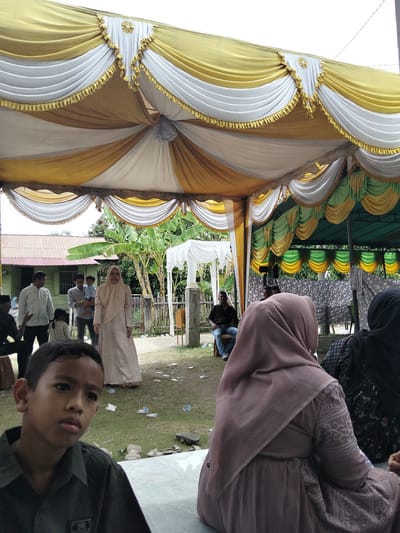 An outdoor event is taking place under a decorative tent. The tent is adorned with yellow and white drapes, featuring gold trim and tassels. The ground is covered with grass and scattered debris, including plastic bottles and paper.
In the foreground, a young boy with dark hair is looking to his left. Behind him, a woman wearing a light brown hijab and dress is seated, her back to the camera. To her right, another woman is partially visible, wearing a black headscarf.
Further back, several other individuals are present. A woman in a light brown hijab stands near a building with a red roof. To her left, a man in a white shirt and a man holding a baby are standing. Another man is seated on a chair to the left of the frame. In the background, there are banana trees and a wooden fence. The setting appears to be a residential area in Cottrueng, Indonesia.