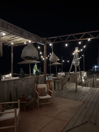 This is a low-angle, full-frame, vertical, outdoor night shot of a rustic, open-air restaurant or bar area in Torreilles, France. The scene is illuminated by string lights and appears to be situated near a beach or water. The foreground features a wooden deck with a textured, rust-colored rug. Two simple wooden chairs with woven seats and patterned cushions are visible. To the left, a small round wooden table holds decorative pottery and a potted plant. Further back, several more wicker chairs and tables are arranged under a thatched-roof canopy. In the background, a disco ball hangs from a wooden tripod, surrounded by more string lights. The overall aesthetic is bohemian and relaxed, with natural materials like wood and straw dominating the scene. The dark sky suggests it is nighttime. There are no people visible in the image.