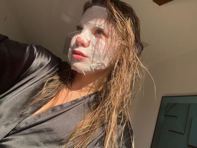 A low-angle, slightly rotated, close-up indoor shot shows a woman wearing a white sheet facial mask, looking at the camera. Her light brown hair is loose and wet, partially obscuring her face. Only her eyes, lips, and nose are visible through the mask. She is wearing a dark-colored, silky robe. A framed artwork with geometric designs hangs on the wall in the background. The location is uncertain. Sotonići, Montenegro is not identifiable in the scene.