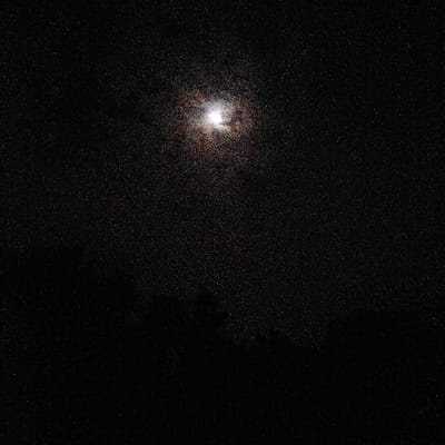 The image displays a dark, nighttime outdoor scene located in Mururi, Kenya. A bright, diffused light source, indicative of the moon, is centered in the upper portion of the frame. The entire field of view is permeated by a high density of small, bright, scattered points. Along the lower horizon, indistinct, dark, amorphous shapes suggest the presence of terrain or vegetation. No individuals, specific objects, actions, or interactions are visible.