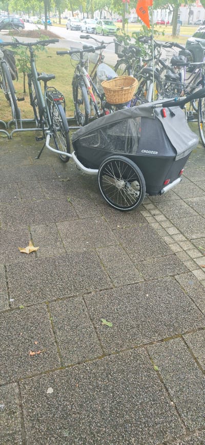 The image shows a bicycle trailer parked on a paved surface in what appears to be a public area, possibly a park or street, with bicycles parked nearby. The bicycle trailer is black with a clear plastic window and a logo that reads "CROOZER". A single fallen leaf is visible on the paving stones in the foreground. In the background, there are several parked bicycles, trees, and a street with cars and buildings. The lighting suggests it is daytime, possibly overcast. Based on the prevalence of bicycles and the urban setting, this scene could be located in many cities. Without further information or contextual clues, it is not possible to definitively identify the specific location within Karlsruhe, Germany.