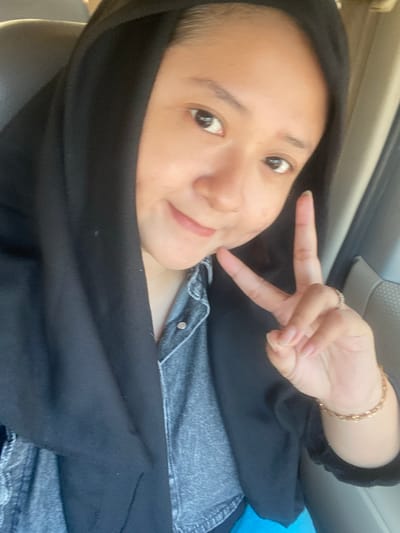 A young woman wearing a black hijab and a denim shirt is inside a vehicle. She is taking a selfie, holding up two fingers in a peace sign with her right hand. A gold bracelet is visible on her wrist. The interior of the car, including a headrest and part of the dashboard, is visible in the background. The image appears to be taken during the daytime, with natural light illuminating the scene.