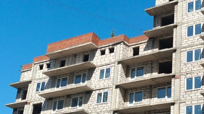 This image shows the exterior of a multi-story building under construction in Zvenigorod, Russia. The building is made of light-colored concrete blocks and has several unfinished floors. Many window openings are visible, some with windows already installed. Concrete balconies or cantilevered floors extend from various levels. The top of the building is capped with red bricks. The sky is a clear, bright blue, and a few thin wires are visible against the sky in the upper left portion of the image. There are no people visible in the frame. The overall impression is of an active construction site.