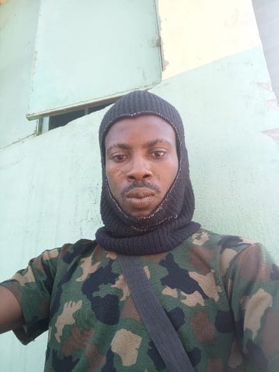 The image depicts an adult male, of apparent Sub-Saharan African descent, positioned in a medium close-up, facing the camera directly. He is wearing a black knitted head covering that functions as a balaclava, exposing his face, and a short-sleeved camouflage patterned shirt. A dark, possibly black, strap is visible diagonally across his chest and left shoulder. The individual has a distinct scar on his left cheekbone and appears to have facial hair, specifically a mustache and chin beard. His expression is neutral. The background consists of a wall painted light green with areas of peeling paint, suggesting deterioration. A rectangular opening or window frame is visible in the upper left portion of the background, and a section of a wall painted light yellow is visible on the upper right. The scene is located in the city of Zaki Biyem, Nigeria.