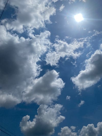 A clear blue sky is visible with scattered white and gray clouds. The sun is brightly shining from the upper right corner, casting a lens flare. In the foreground, the tops of power lines are visible. This scene is located in Berangas Timur, Indonesia.