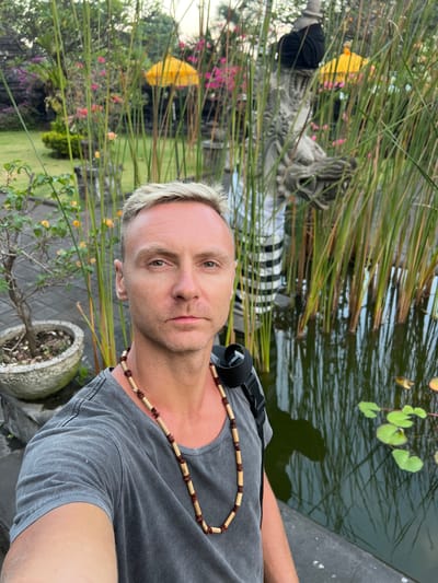 In the city of Dawan, Indonesia, a man with short, bleached blonde hair and a serious expression takes a selfie in front of a tranquil pond. He wears a grey t-shirt and a wooden beaded necklace. Behind him, tall reeds rise from the water, their reflections visible on the dark surface. In the background, hints of a traditional Balinese temple can be seen, including a stone statue and a yellow umbrella. The scene suggests a moment of quiet contemplation in a culturally rich and natural setting.