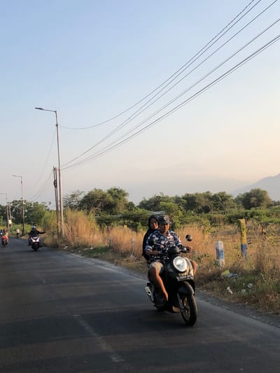 The image depicts a rural roadside scene in Glagah Arum, Indonesia, bathed in the warm light of what appears to be late afternoon or early morning. The foreground is dominated by a dark-colored scooter being ridden by a man wearing a patterned shirt and a hat, with a passenger, a woman in a dark headscarf, seated behind him. The scooter has a license plate that reads "W 4029 NAZ". They are traveling on a paved road with a faint white line marking the lane.
To the left of the road, several other motorbikes are visible further in the distance, also traveling in the same direction. The roadside is characterized by dry, tall grass and sparse vegetation, with a few scattered trees and bushes forming a natural backdrop. Several utility poles with overhead power lines are prominent features along the road. In the far distance, hazy mountains can be seen against a light-colored sky. The overall impression is of a tranquil, everyday scene in a semi-rural Indonesian setting.