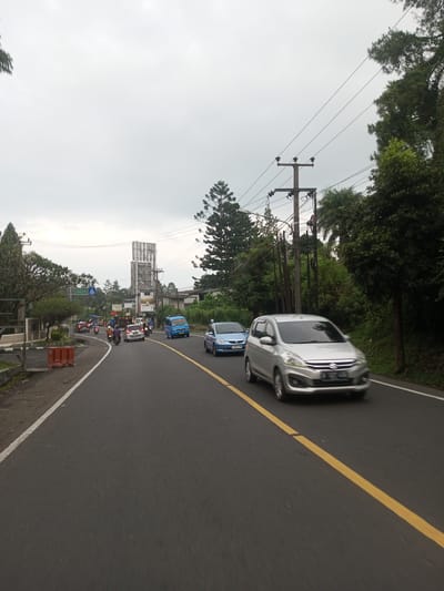 The image shows a road scene with traffic. A silver car is in the foreground, moving along the right side of the road, with a yellow line marking the lane. A light blue car and a blue minivan are behind it. There are several motorcycles further back in the scene. The road is paved, and there are trees on both sides. Utility poles and wires are visible along the roadside. The sky is overcast. A large metal structure is visible in the distance. The location is not Taunggyi, Myanmar.