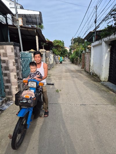 An outdoor scene in Hội An Đông Ward, Vietnam, depicts a paved, narrow residential street under a clear, bright sky. In the foreground, an adult male, appearing middle-aged or elderly, wears a white tank top, khaki pants, and black sandals. He is seated on a blue motor scooter, holding a young child, dressed in a patterned t-shirt, on the handlebars in front of him. The man smiles towards the camera; the child looks slightly off-camera. The scooter has a black wire basket mounted on the handlebars, containing a light-colored cap and other items. To the left of the scooter, a low wall with grey and brown patterned tiles and a turquoise metal gate are visible. The concrete street, marked by cracks and a central seam, recedes into the distance, lined by residential buildings and dense green foliage. On the right, a weathered white wall with green discoloration and a black metal gate stands opposite the subjects. Overhead, numerous electrical or communication wires crisscross the street, supported by a prominent utility pole. In the distant midground, a person on a two-wheeled vehicle carrying a large green object travels away from the viewer.