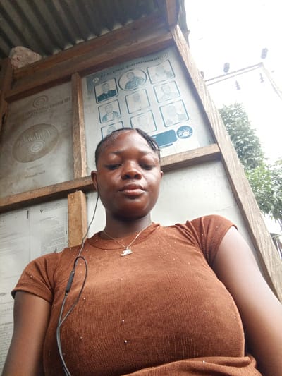 A young, dark-skinned woman with braided hair stands in Mbribit, Nigeria. She is wearing a brown ribbed t-shirt with subtle sparkling accents and a necklace with a small pendant. Earphones are visible, plugged into her ear. She appears to be in a covered outdoor area with wooden supports and corrugated metal roofing. Behind her is a framed bulletin board with posters and printed documents. Trees are visible in the background.