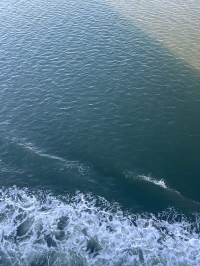 This image depicts a dynamic body of water, likely a river or estuary, with two distinct water types meeting. The lower left of the frame is dominated by churning white foam and waves, indicative of a moving vessel's wake. The water transitions to a deep blue-green hue. The upper right portion of the image shows a contrasting lighter, murkier water, suggesting a confluence with another water source, possibly carrying sediment or having a different temperature. The surface of the water exhibits a gentle ripple pattern throughout, catching ambient light. There are no visible people, structures, or specific landmarks that would allow for the identification of the location within Batas Desa Skotong Timur, Indonesia. The scene is an abstract representation of water dynamics.