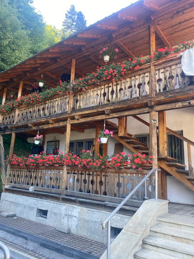 The image depicts a rustic wooden building with multiple levels of balconies adorned with abundant red and pink flowers in planters. The architecture is characterized by exposed wooden beams and decorative wooden railings with circular and cross-shaped cutouts. An outdoor staircase, also made of wood, ascends the side of the building, featuring a metal handrail. The building appears to be situated in a natural, possibly mountainous, environment, with lush green trees visible in the background. On one of the lower balconies, a framed religious icon is hung on the wall next to a window. The ground level features a concrete foundation with metal grating and stone elements. A small, rectangular window is visible in the concrete foundation. The scene suggests a serene and picturesque setting, likely in a rural area or a mountain resort.