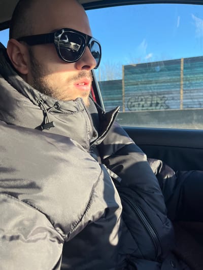 A male individual is depicted from the chest up, seated in the passenger side of a vehicle in Saint-André-lez-Lille, France, under clear daylight conditions. He has short hair, stubble, and wears black sunglasses and a black puffer jacket with a partially visible zipper and a black drawstring toggle. His head is turned towards the right, with his mouth slightly open. Strong sunlight illuminates the left side of his face and jacket, casting shadows. Through the vehicle's window, a clear blue sky is visible above a roadside barrier constructed of horizontal panels, which appear light blue or greenish-blue. Black graffiti spelling "GREX" is present on a lower panel of this barrier. Bare trees are visible in the distance beyond the barrier. The interior of the vehicle shows a red car door frame.