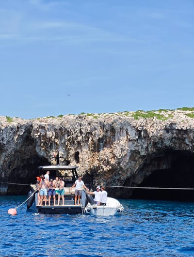In the city of Grad Vis, Croatia, on a bright, clear day with a blue sky, a group of people are enjoying a boat trip near a rocky cliff face with visible caves. Two boats are present in the vibrant blue water. The larger boat, a dark-colored vessel with a canopy, carries several individuals, some of whom are standing and taking photos. A red buoy floats in the water near this boat, tethered by a rope. A smaller, white boat is positioned alongside the larger vessel, with two people on board, one wearing a white shirt with lettering that appears to read "CAPTAIN CREW". The overall scene depicts a leisurely outing with people enjoying the coastal scenery.
